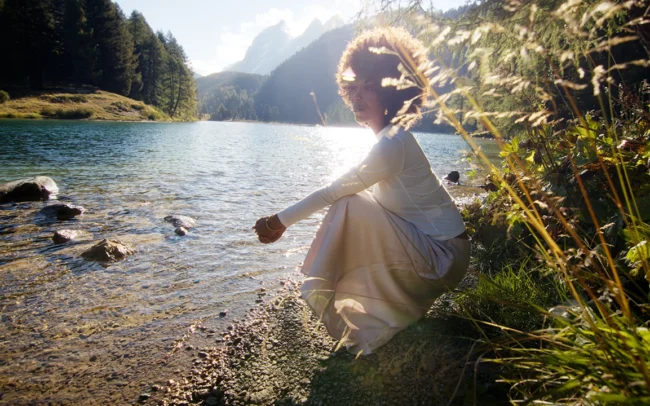 A woman with an afro dressed in white sitting by a lake in the Swiss Alps. The sun is shining, everything is glowing.