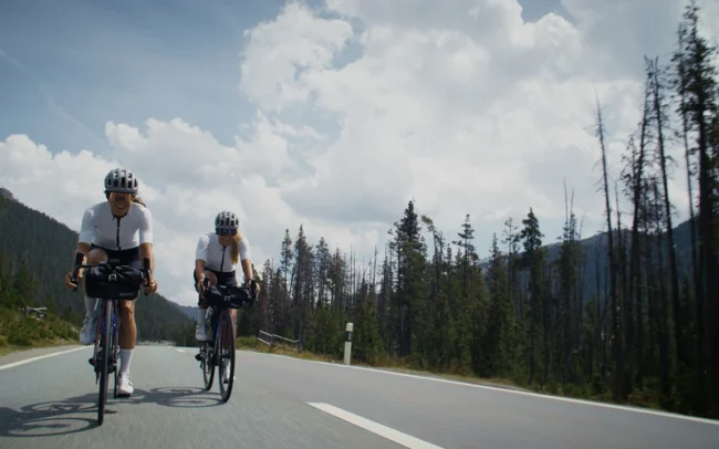 Magdalean Mittersteiner and Elisa Deutschmann cycling with a smile on a paved road in sunshine.