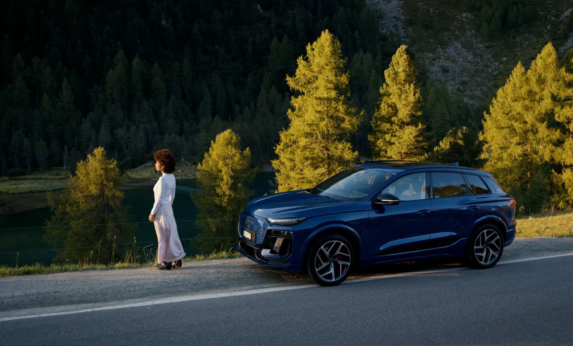 A womand standing next to her AUDI Q6 e-tron in front of beautiful colored autumn trees.