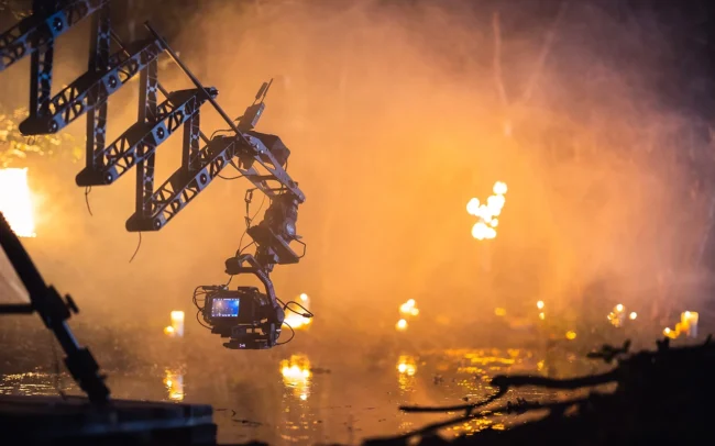 A camera crane is pushing over a water surface in a dark forest lit by flames.