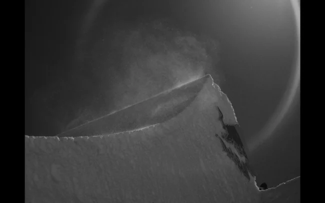 A kicker seen from a low angle. There are snow particles in the air. The scene is black and white.