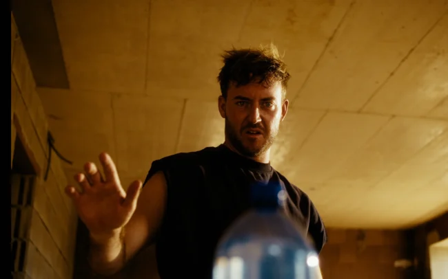 Dramatic close-up of a construction worker hesitantly reaching out toward a mystery water bottle in the Austrian Kiwi comedy sketch.