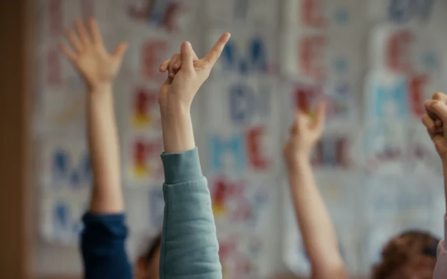 Close-up shot of students enthusiastically raising their hands in a classroom to answer a question during the English lesson sketch by Holy Moly films.